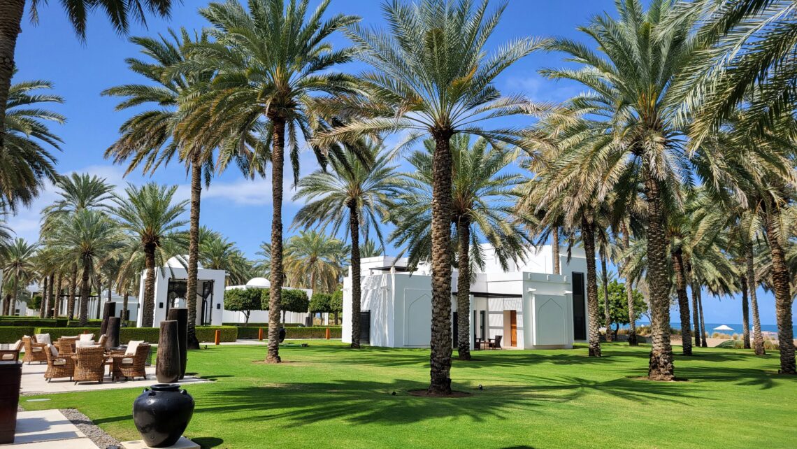 Garden of the Chedi Hotel in Muscat Oman, featuring Palmtrees, lucious green grass and the ocean in the background. You can also see the white and black arabian style buildings and a terrace with wooden stools.