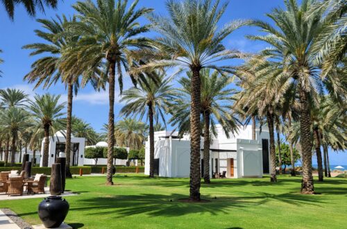 Garden of the Chedi Hotel in Muscat Oman, featuring Palmtrees, lucious green grass and the ocean in the background. You can also see the white and black arabian style buildings and a terrace with wooden stools.