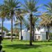 Garden of the Chedi Hotel in Muscat Oman, featuring Palmtrees, lucious green grass and the ocean in the background. You can also see the white and black arabian style buildings and a terrace with wooden stools.